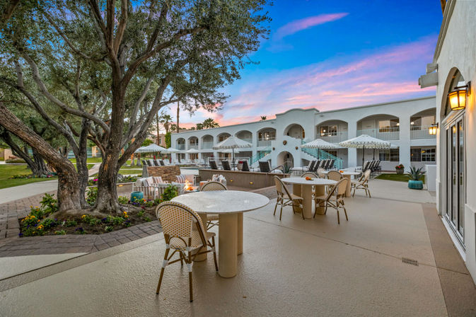 Inviting Mediterranean-style hotel courtyard at sunset with round outdoor dining tables and wicker chairs, mature olive trees, poolside loungers and umbrellas in front of a two-story white arched building