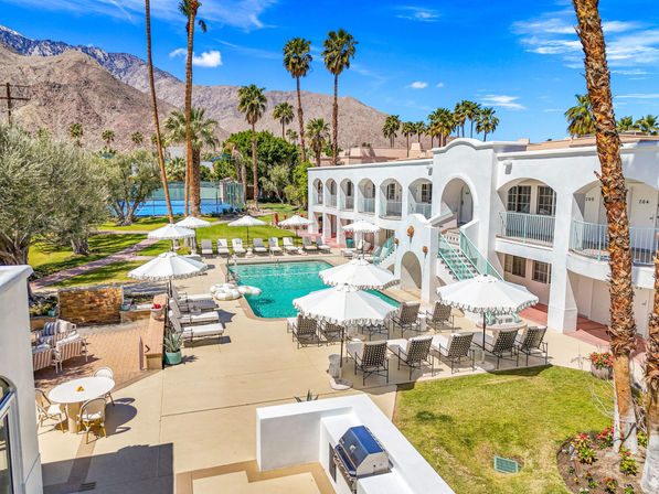 Sunny desert resort courtyard with white stucco two-story hotel, turquoise swimming pool lined with lounge chairs and parasols, tall palm trees, tennis courts, and rocky mountain backdrop