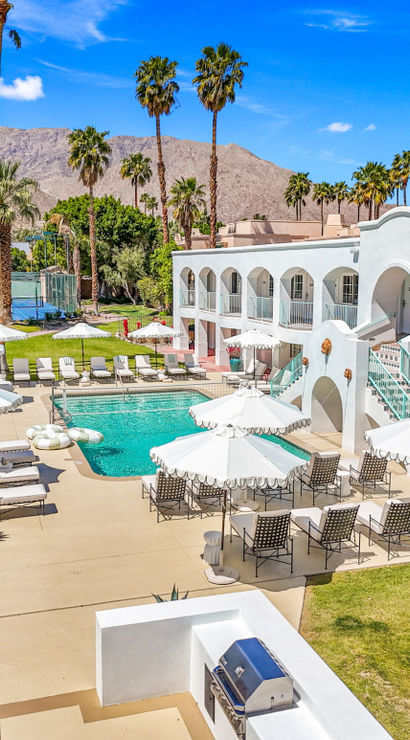 Sunny desert resort courtyard with white stucco two-story hotel, turquoise swimming pool lined with lounge chairs and parasols, tall palm trees, tennis courts, and rocky mountain backdrop