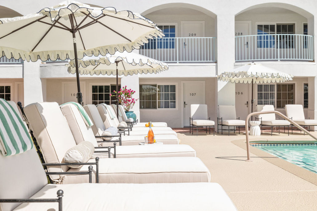 Sunlit hotel pool deck with white cushioned chaise lounges, scalloped umbrellas, striped towels and balcony rooms overlooking a turquoise pool.