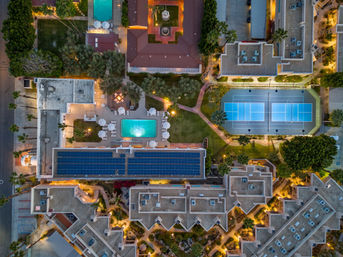 Aerial evening drone view of a resort complex with illuminated swimming pool, blue tennis courts, rooftop solar panels, palm trees and winding pathways.