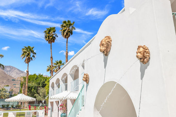 White stucco courtyard with bronze lion-head water spouts, pastel railings, tall palm trees and desert mountains under a bright blue sky