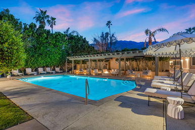 Glowing blue outdoor resort swimming pool at dusk, surrounded by lounge chairs, umbrella-shaded cabanas, palm trees and a mountain backdrop.