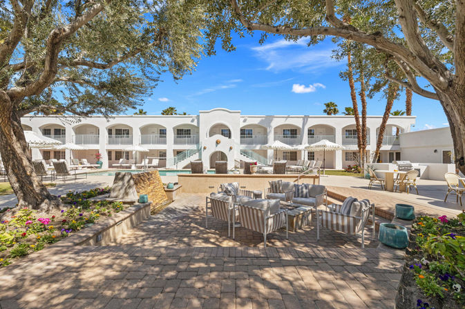 Sunlit desert resort courtyard with a white two-story Mediterranean-style hotel, central swimming pool, striped patio lounge seating, shaded seating under mature trees and tall palm trees against a bright blue sky.
