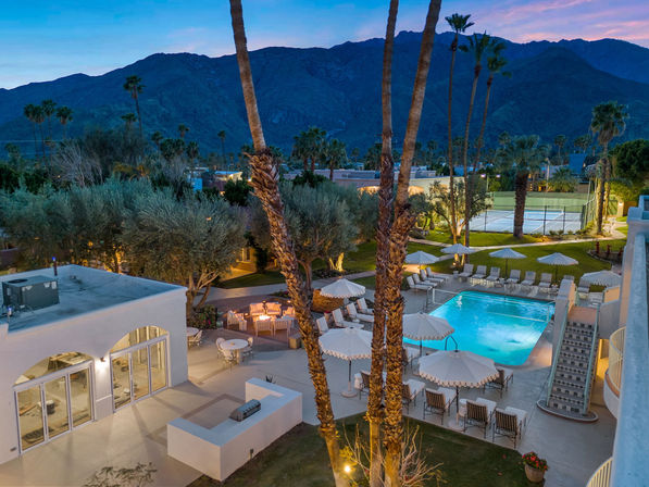 Twilight aerial view of a desert resort courtyard with an illuminated rectangular pool, tall palm trees, poolside umbrellas and lounge chairs, outdoor dining seating and nearby tennis courts set against rugged mountains.