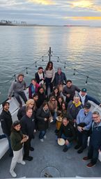 Group of about 20 people smiling on a boat deck under string lights, enjoying food and drinks at sunset over a calm coastal bay with a palm-lined shoreline and pier in the distance.