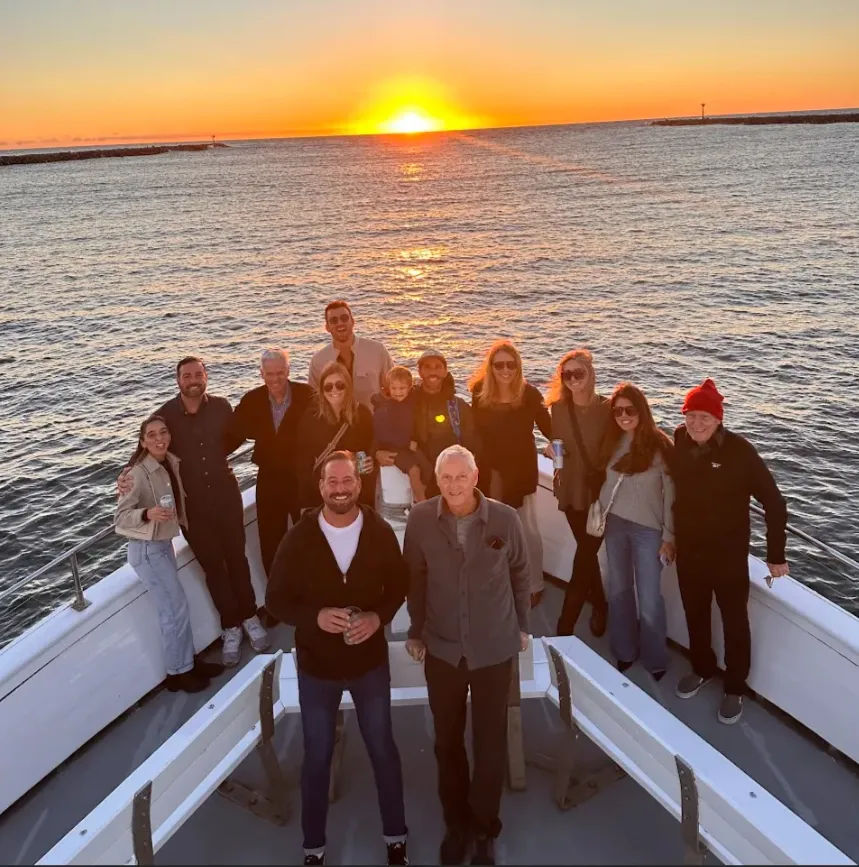 Group of people posing on the bow of a boat at golden sunset over the ocean, smiling and relaxed with a child and a guest in a red beanie, sun reflecting on the water.