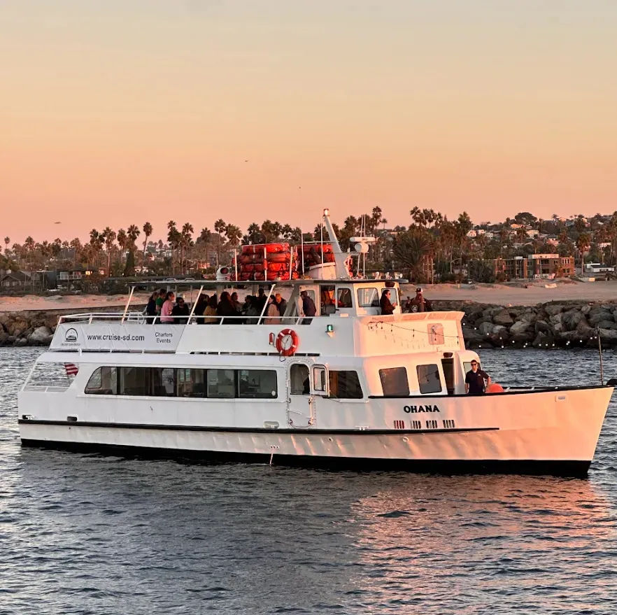 Sunset sightseeing boat with passengers gliding past a palm‑lined shoreline and rocky breakwater, warm orange sky reflecting on calm harbor water.