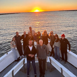 Sunset cruise: smiling group gathered on the bow of a boat as a golden sun dips below the ocean with harbor breakwaters on the horizon