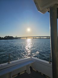 Boat deck railing in foreground, sun low over a coastal waterway with sparkling reflections, a bridge span and palm-tree silhouettes on the horizon.