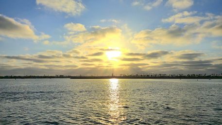 Serene coastal sunset with golden sun reflecting across rippled bay waters toward a distant palm-lined shoreline under scattered clouds.