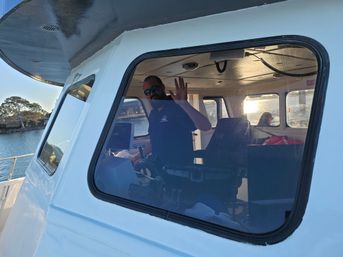 Crew member waving from a boat wheelhouse seen through a cabin window, helm and navigation gear inside, sunny harbor shoreline in the background.