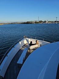 Bow of a white boat cruising a sunny coastal harbor with two people seated on a front bench, calm blue water, distant marina and palm-lined shoreline under a clear sky