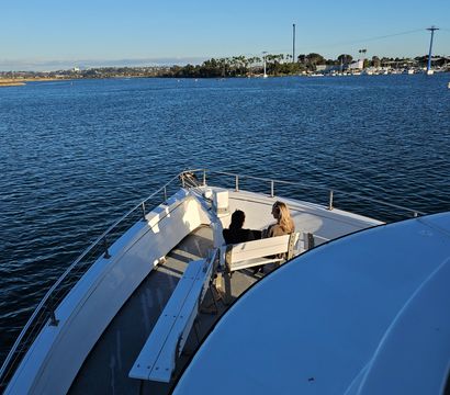 Bow of a white boat cruising a sunny coastal harbor with two people seated on a front bench, calm blue water, distant marina and palm-lined shoreline under a clear sky