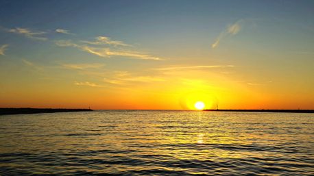 Golden sunset over a calm ocean, sun sinking to the horizon with shimmering rippled water and a silhouetted breakwater