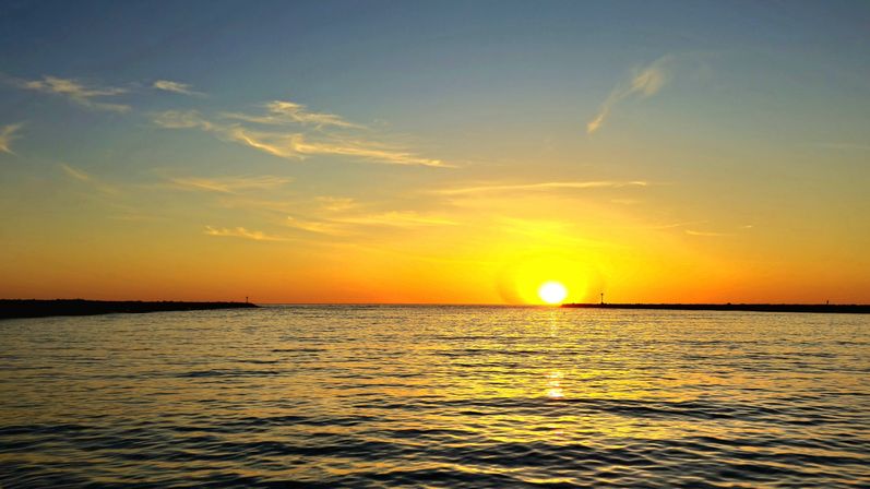Golden sunset over a calm ocean, sun sinking to the horizon with shimmering rippled water and a silhouetted breakwater