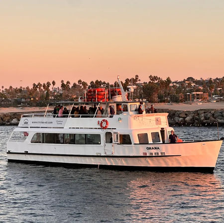White two-deck passenger yacht with people on deck cruising along a palm-lined California coastline at golden sunset, harbor waters reflecting warm light.