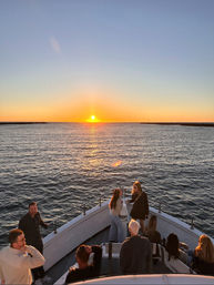 Sunset cruise scene: people on a boat bow watching the golden sun sink over calm ocean waters with its reflection leading to distant harbor breakwaters.