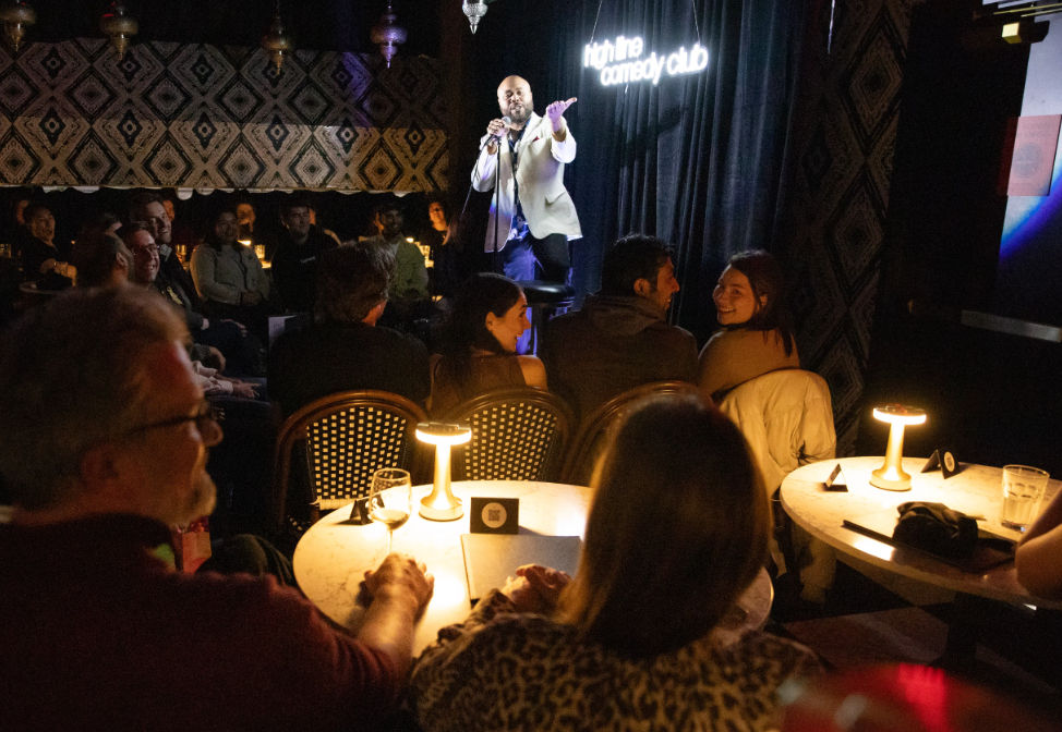 Intimate downtown comedy club at night — a stand-up comedian on stage entertaining a laughing, candlelit audience seated at small tables.