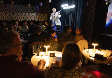 Intimate downtown comedy club at night — a stand-up comedian on stage entertaining a laughing, candlelit audience seated at small tables.