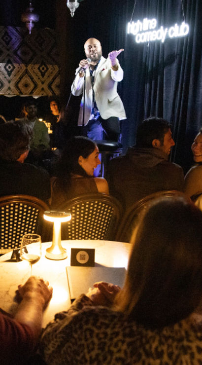 Intimate downtown comedy club at night — a stand-up comedian on stage entertaining a laughing, candlelit audience seated at small tables.