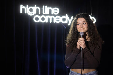 Curly-haired stand-up comedian smiling and holding a microphone on a small comedy club stage with a glowing neon comedy sign and dark velvet curtains
