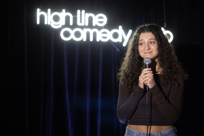 Curly-haired stand-up comedian smiling and holding a microphone on a small comedy club stage with a glowing neon comedy sign and dark velvet curtains