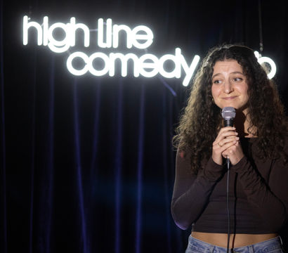 Curly-haired stand-up comedian smiling and holding a microphone on a small comedy club stage with a glowing neon comedy sign and dark velvet curtains