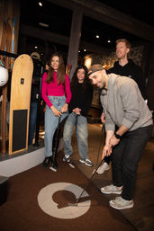 Four friends playing indoor mini-golf in a cozy ski-lodge style venue; a man lines up a putt while three friends watch near vintage skis and helmets.