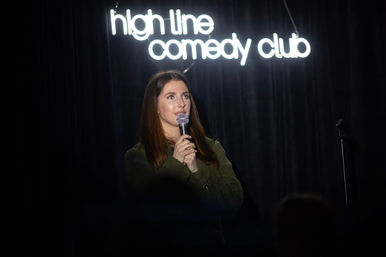 Woman stand-up comedian holding a microphone on a small comedy club stage beneath a glowing neon sign, dark curtain backdrop and audience silhouettes.