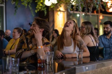 Group of young adults laughing at a lively indoor bar counter with cocktails, hanging greenery and warm ambient lighting — upbeat nightlife scene