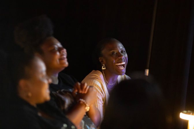 Three audience members laughing in a dimly lit live comedy club, warm spotlight on a smiling person seated near a microphone stand.