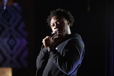 Stand-up comedian in a black hoodie holding a microphone and touching his neck, performing under a spotlight on a dim comedy club stage.