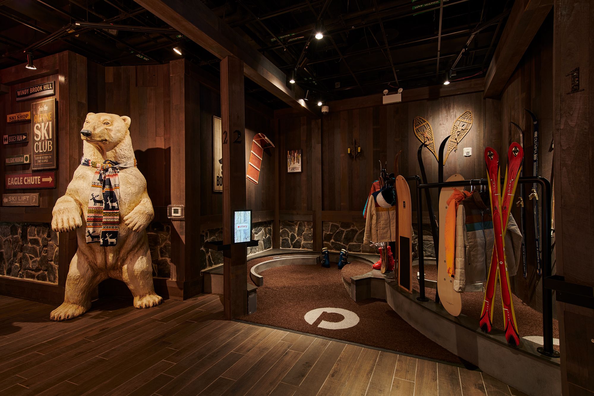 Playful carved polar bear wearing a patterned scarf in a warm wooden ski-lodge boot room with red skis, snowshoes, a snowboard, jackets and ski boots.