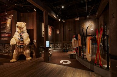 Playful carved polar bear wearing a patterned scarf in a warm wooden ski-lodge boot room with red skis, snowshoes, a snowboard, jackets and ski boots.