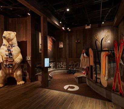 Playful carved polar bear wearing a patterned scarf in a warm wooden ski-lodge boot room with red skis, snowshoes, a snowboard, jackets and ski boots.