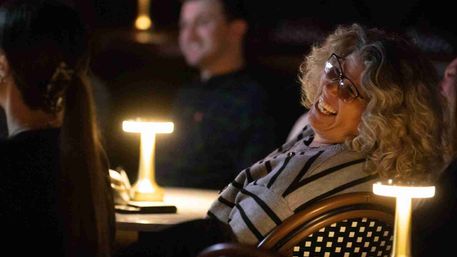 Curly-haired woman in glasses laughing while seated in a cozy, dimly lit venue at a table with warm glowing lamps and other audience members in the background.