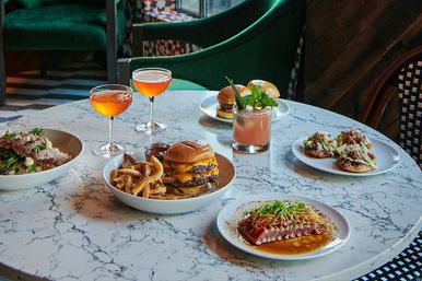 Marble-top restaurant table with a vibrant spread: double cheeseburger and fries, seared tuna with microgreens, small tostadas, a steak rice bowl, two orange cocktails and a pink mint cocktail.