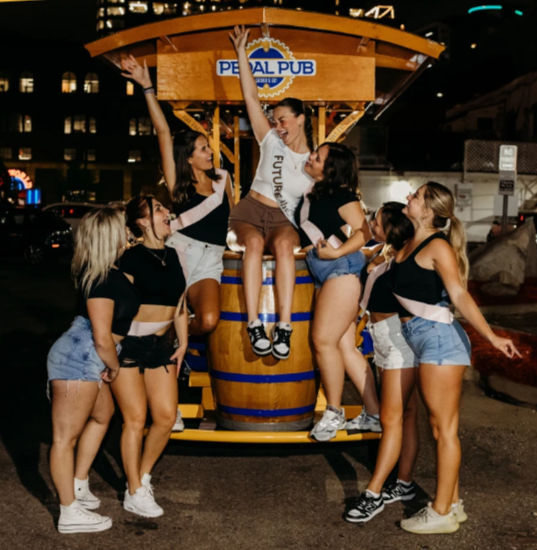 Bachelorette group of friends wearing sashes laughing and posing around a wooden barrel on a pedal-powered party bar at night on a city street.
