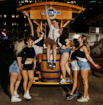 Bachelorette group of friends wearing sashes laughing and posing around a wooden barrel on a pedal-powered party bar at night on a city street.