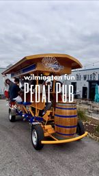 Group riding a yellow pedal-pub bike in Wilmington with a wooden barrel at the front, outdoor patio seating and cloudy sky