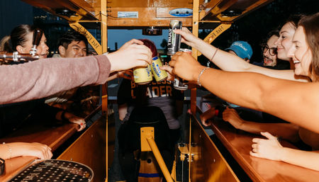 Group of friends laughing and toasting canned drinks aboard a yellow party bike/pedal pub at dusk on a city street