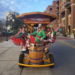 Women in festive elf costumes riding a yellow pedal-powered party bike with a central wooden barrel on a downtown city street lined with brick apartment buildings.