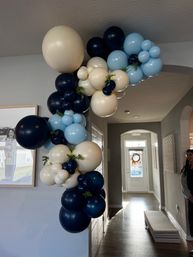 Indoor balloon garland framing a home entryway: mixed-size navy, light-blue and cream balloons with small green foliage accents decorating an arched hallway near the front door.
