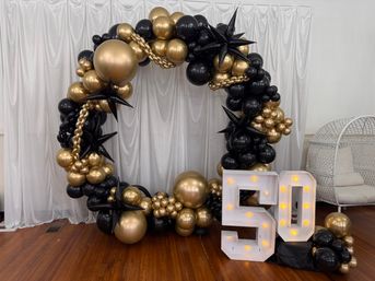 Indoor black-and-gold balloon arch with star-shaped balloons and gold chain accents framing illuminated marquee "50" — glam 50th birthday party backdrop on wood floor