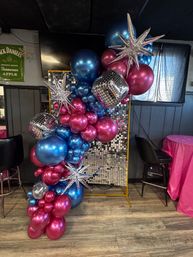 Indoor event photo backdrop with cascading metallic blue and fuchsia balloons, disco mirror balls and silver starburst accents against a shimmering sequin panel