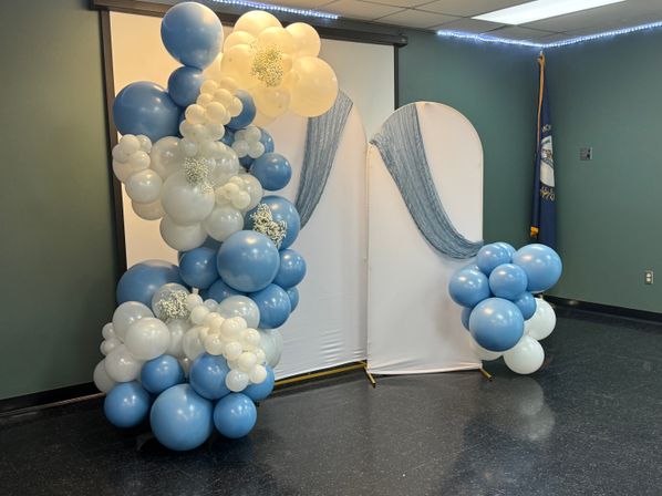 Indoor photo backdrop in a community room with cascading blue and white balloon garlands, clusters of baby’s-breath florals and a sheer blue drape for a baby shower or celebration.