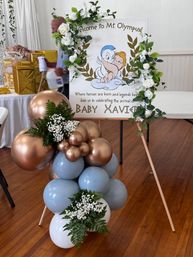 Indoor baby shower display with 'Mt. Olympus' cherub welcome sign framed by white rose garland, rose-gold and pale blue balloons, and greenery on a wooden floor.