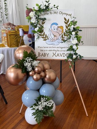 Indoor baby shower display with 'Mt. Olympus' cherub welcome sign framed by white rose garland, rose-gold and pale blue balloons, and greenery on a wooden floor.