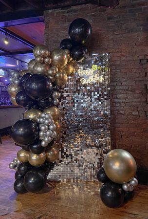 Black and gold balloon garland framing a mirrored sequin photo backdrop with neon "Happy Birthday" sign, set against an exposed brick wall in a cozy indoor bar/restaurant celebration setup.